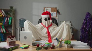 İşte hayalet gibi giyinmiş biri. Beyaz çarşaflı cadılar bayramı işadamı hayaleti ve Noel Baba şapkası masasında oturuyor, Noel için süslenmiş koltukta mutlu bir şekilde dans ediyor. Cadılar Bayramı iş konseptiniz kutlu olsun..