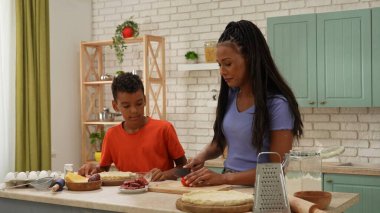 Afro-Amerikalı mutlu aile mutfakta yemek hazırlıyor. Oğlum, annenin pizza için domates dilimleyişini izliyor. Birlikte vakit geçirip, sağlıklı bir hayat sürüyoruz..