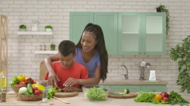 Afro-Amerikalı mutlu aile mutfakta yemek hazırlıyor. Annem salata için salatalık doğramaya yardım ediyor. Birlikte zaman geçirmek, sağlıklı bir yaşam tarzı..