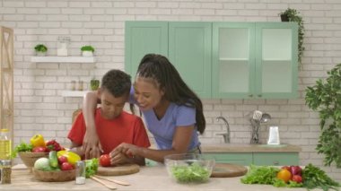 Afro-Amerikalı mutlu aile mutfakta yemek hazırlıyor. Annem salata için domates doğramaya yardım ediyor. Birlikte zaman geçirmek, sağlıklı bir yaşam tarzı..