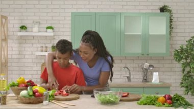Afro-Amerikalı mutlu aile mutfakta yemek hazırlıyor. Annem salata için domates doğramaya yardım ediyor. Birlikte zaman geçirmek, sağlıklı bir yaşam tarzı..