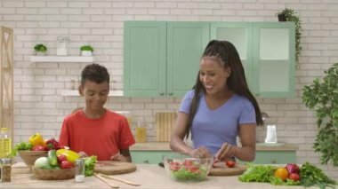 Afro-Amerikalı mutlu aile mutfakta yemek hazırlıyor. Annem salata için domates doğramaya yardım ediyor. Birlikte zaman geçirmek, sağlıklı bir yaşam tarzı..
