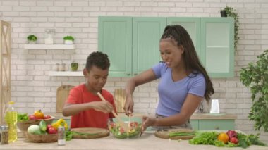 Afro-Amerikalı mutlu aile mutfakta yemek hazırlıyor. Anne ve oğlu salatayı neşeyle ahşap kaşıklarla karıştırıyorlar. Birlikte zaman geçirmek, sağlıklı bir yaşam tarzı..