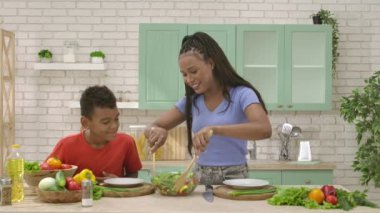 Afro-Amerikalı mutlu aile mutfakta yemek hazırlıyor. Annem tahta kaşıklarla tabağa salata koyar. Birlikte zaman geçirmek, sağlıklı bir yaşam tarzı..
