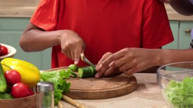 Afro-Amerikalı mutlu aile mutfakta yemek hazırlıyor. Annem salata için salatalık doğramaya yardım ediyor. Birlikte zaman geçirmek, sağlıklı bir yaşam tarzı..