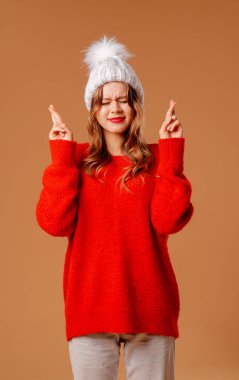 Kırmızı kazaklı ve şapkalı Kar Kızı bej arka planda stüdyoda poz veriyor. Genç kız parmaklarını çapraz tutuyor. Yeni yıl ve Noel tatili konsepti.