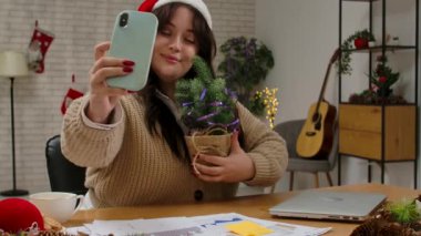 Tatil sezonunda evden çalışırım. Noel Baba şapkalı genç bir iş kadını yeni yıl ofis dekorasyonunda dizüstü bilgisayarda çalışıyor, akıllı telefonuyla selfie çekiyor, elinde şenlikli ağaçla gülümsüyor..