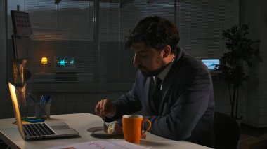 Odaklanmış profesyonel erkek, geç saatlere kadar çalışıyor. Yetişkin bir işletme yöneticisi, geceleri ofiste dizüstü bilgisayar başında oturup çay fincanıyla kek yiyor..