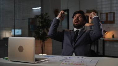 Odaklanmış profesyonel erkek, geç saatlere kadar çalışıyor. Yetişkin iş adamı menajeri masada oturup bilgisayarla çalışıyor, akıllı telefonla el ele tutuşuyor, mutlu bir gülümseme, kazanan jestler..