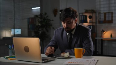Odaklanmış profesyonel erkek, geç saatlere kadar çalışıyor. Yetişkin bir işletme yöneticisi masa başında oturuyor, akşam ofiste dizüstü bilgisayarla çalışıyor, bir fincan çayla bir parça kek yiyor ve yemek yiyor..