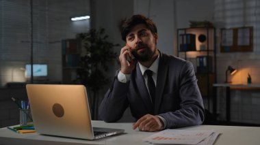 Odaklanmış profesyonel erkek, geç saatlere kadar çalışıyor. Yetişkin iş adamı yöneticisi masada oturuyor, akşam ofiste dizüstü bilgisayarla çalışıyor, telefonlara cevap veriyor, akıllı telefonla konuşuyor, olumlu ifade veriyor..