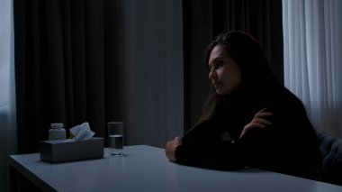 Karanlık dairede stresli bir kadın. Akıl sağlığıyla ilgili sorunları olan bir kadın, kameranın önünde masada oturuyor, depresif bir yüz ifadesi, ilgisiz bir yüz ifadesi, yan görüş..