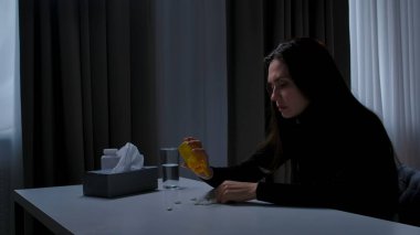 Karanlık dairede stresli bir kadın. Akıl sağlığıyla ilgili sorunları olan bir kadın, kameranın önünde masada oturuyor, depresif bir ifade, masaya bir kavanoz dolusu hap dök, yan görüş..