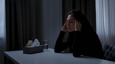 Karanlık dairede stresli bir kadın. Akıl sağlığı sorunları olan bir kadın, kameranın önünde masada oturuyor, depresif bir ifade, masaj, baş ağrısı, yan görüş....