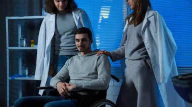 Sick male patient resting in medical clinic ward. Adult man patient in hospital ready for discharge sitting in wheelchair and talking, friends supporting man smiling positive, middle shot. clipart