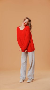 Kış kıyafetli gülümseyen kadın kameraya poz veriyor. Atkuyruklu neşeli bir genç kadın, kırmızı kazak ve gri kot pantolon giyiyor. Turuncu stüdyo arka planında izole edilmiş şirin poz veriyor ve güzel görünüyor..