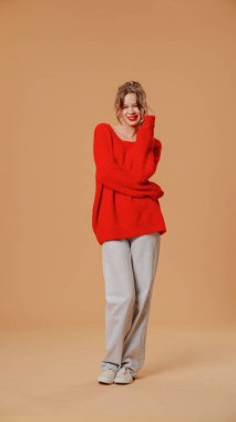 Kış kıyafetli gülümseyen kadın kameraya poz veriyor. Atkuyruklu neşeli bir genç kadın, kırmızı kazak ve gri kot giyiyor, kameraya gülümsüyor, turuncu stüdyo arka planında izole edilmiş..