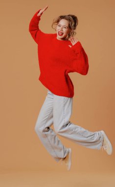 Kış kıyafetli gülümseyen kadın kameraya poz veriyor. Kırmızı kazaklı ve gri pantolonlu neşeli genç bir kadın kameraya mutlu atlama pozuyla poz veriyor, neşeli gülümsüyor, turuncu stüdyo arka planında izole edilmiş..