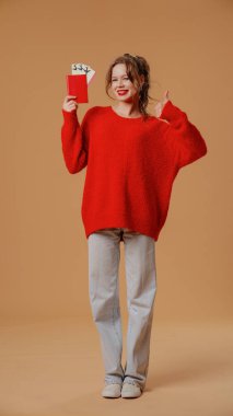 Smiling female in winter outfit poses at camera. Cheerful young woman in red jumper and grey jeans posing with flight tickets, smiling shows thumb up like gesture, isolated on orange studio background clipart