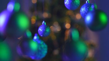 Yeni yıl dekorasyonları. Yeşil mavi neon ışıkta Noel ağacı topları, yukarıdan sarkan parlak renkli oyuncaklar, ışıltılı süsler yakın çekim, bulanık arka plan şenliği..