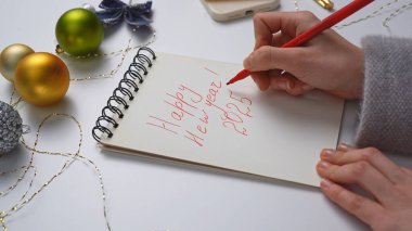 Yeni yıl kararı ve dilek listesi. Noel 'in yakın çekimleri, dekore edilmiş beyaz masa, dizüstü bilgisayarlı kadın el yazısı, sarmal defterli yeni yıl tebrikleri, üst görünüm..