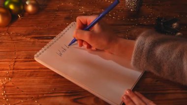 Yeni yıl kararı ve dilek listesi. Noel 'in yakın çekimleri, dekore edilmiş ahşap masa, not defterine mavi kalemle yazan kadın, dilek listesi, aşk, para ve mutluluk diliyor..