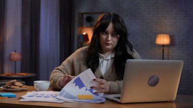 Evden uzakta bağımsız bir iş. Genç iş kadını masa başında oturuyor, geç saatlere kadar ofis kumandasında çalışıyor dizüstü bilgisayara bakıyor, elinde belgelerle arama yapıyor..