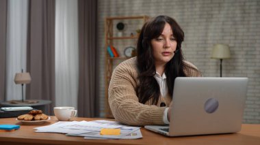 Evden uzakta bağımsız bir iş. Genç iş kadını serbest çalışan masa başında oturuyor ev ofisinin kumandasında çalışıyor, kulaklık takıyor, bilgisayardaki video konuşmalarıyla konuşuyor, odaklanmış ifade.