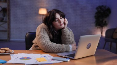 Evden uzakta bağımsız bir iş. Genç iş kadını masa başında oturuyor, geç saatlere kadar ev ofisinin kumandasında çalışıyor, dizüstü bilgisayar ekranına bakıyor, uykulu yorgun bir ifade takınıyor, başını elinde tutuyor..