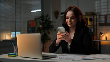 Yetişkin bölük lideri fazla mesai yapıyor. Genç iş kadını geç saatlere kadar masa başında çalışır, dizüstü bilgisayar kullanır, akıllı telefonda yazı yazar, gülümseyen bir ifade, orta çekim yapar..