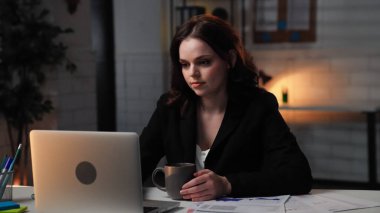 Ofiste çalışan bir iş adamı. Genç iş kadını geç saatlere kadar masa başında çalışır, dizüstü bilgisayar kullanır, ekrana bakar, elinde kahve fincanı, odaklanmış pozitiflik, yan çekim....