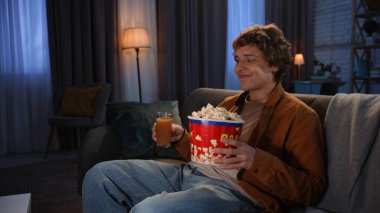 Adam evde boş vakit geçiriyor. Genç adam akşamını evde, oturma odasında kanepede oturup, televizyon izleyerek, elinde patlamış mısır ve meyve suyu tutarak, iştahla yiyeceğe bakarak, olumlu bir ifadeyle..