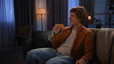 Adam evde boş vakit geçiriyor. Genç bir adam akşamını evinde, oturma odasındaki koltukta oturup elinde altın kredi kartı tutarak mutlu bir şekilde gülümseyerek geçirir..