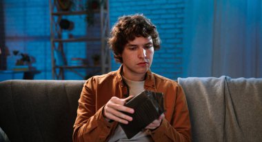 Adam evde boş vakit geçiriyor. Genç bir adam akşamını evde kanepede oturup cüzdanını tutarak ve ciddi odaklanmış yüz ifadesiyle içeri bakarak geçiriyor..