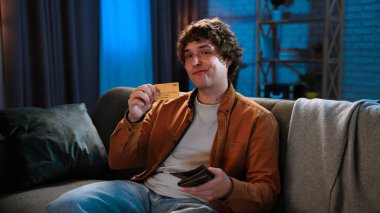 Adam evde boş vakit geçiriyor. Genç bir adam akşamını evinde, oturma odasındaki koltukta oturup elinde altın kredi kartı tutarak mutlu bir şekilde gülümseyerek geçirir..