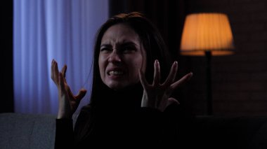 Karanlık dairede depresif bir kadın. Ruhsal sorunları olan bir kadın silueti, koltukta oturuyor, depresif bir ifade, sinirli bir ağlayan yüz, el ele tutuşuyor, yakın çekim..