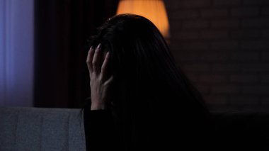 Karanlık dairede depresif bir kadın. Ruhsal sorunları olan bir kadın silueti, koltukta oturuyor, depresif bir ifade, elleri kafasından tutarak yüzünü kapatıyor, yakın çekim..