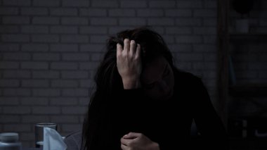 Karanlık dairede stresli bir kadın. Akıl sağlığıyla ilgili sorunları olan bir kadının silüeti masada oturuyor, depresif bir ifade takınıyor, elleriyle başını tutuyor, yüzünü gizliyor, yakın çekim yapıyor..