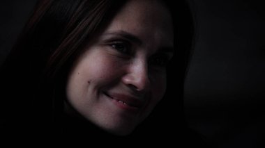 Karanlık dairede depresif bir kadın. Akıl sağlığıyla ilgili sorunları olan bir kadının silueti, kameranın önünde masada oturuyor, depresif bir ifade, düşünüyor, acı bir gülümseme, yakın çekim..