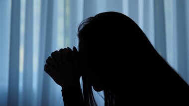 Karanlık dairede stresli bir kadın. Zihinsel sağlık sorunları olan kadın silueti, pencerenin önünde oturuyor, depresif ifade, aşağıya bakıyor, düşüncelere dalmış, melankolik kayıtsız yüz, kenar çekim.