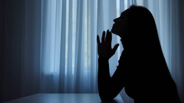 Karanlık dairede stresli bir kadın. Zihinsel sağlık sorunları olan kadın silueti, pencerenin önünde oturmak, depresif ifade, dokunaklı çene düşüncesi, melankolik ilgisiz yüz, kenar çekim.