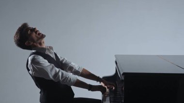 Klasik piyano konseri. Piyanist siyah piyanonun üzerinde oturuyor ve konser sahnesinde etkileyici bir şekilde şarkı söylüyor, yakın çekim gri stüdyo arka planında izole edilmiş..