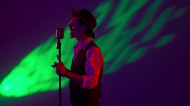 Konser salonunda sahne alan bir besteci. Şık şapkalı adam, karanlık konser sahnesinde kırmızı neon ışıkta duruyor ve renkli stüdyo arka planında izole edilmiş retro mikrofonla şarkı söylüyor..