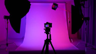 Profesyonel bir stüdyonun içinde. Modern ışıklandırma ekipmanlarıyla fotoğraf ve video çekimi için profesyonel stüdyo, Pembe neon ışıkla renklendirilmiş siklonama arkaplan, tripodun üzerindeki kamera.