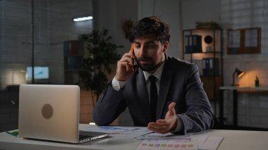 Odaklanmış profesyonel erkek, geç saatlere kadar çalışıyor. Yetişkin bir işletme yöneticisi, masa başında oturuyor, akşam ofiste fazla mesai yapıyor, bilgisayar başında arama yapıyor, akıllı telefondan sinirli bir şekilde konuşuyor..