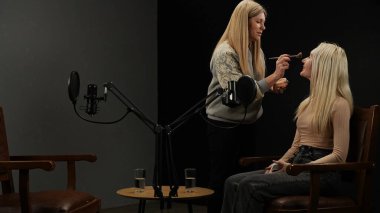 Çevrimiçi podcast gösterisi. Podcast stüdyosundaki şık deri koltukta oturan bayan mikrofonlar, kayıt için hazırlanan röportaj, stilist makyajı ve görünüşünü düzeltmeye geliyor..