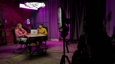 Çevrimiçi yayın. Erkek sunucu ve kadın konuk kayıt stüdyosunda mikrofon kullanarak röportaj yaparken sahne arkası görüntüsü. Erkek ve kadın talk show 'u başlatırken kameraman yan tarafta oturuyor..