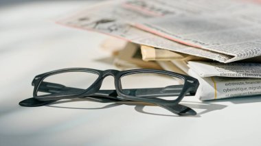 Bir gazeteye yakın plan gözlük. Masanın üzerinde siyah çerçeveli gözlüklerle duran bir gazeteyi gösteren bir fotoğraf..