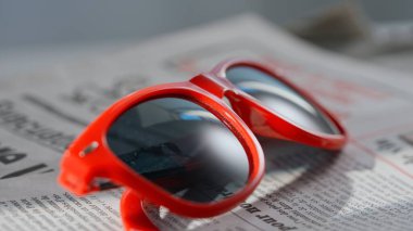 A close up of the glasses on the newspaper. Shot showing of a newspaper lying on a table with red square-rimmed sunglasses on it. clipart