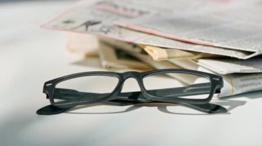 Gazetedeki gözlüklerin yakın çekimi. Üzerinde siyah kare çerçeveli gözlüklerle masanın üzerinde duran bir gazetenin hareketli bir görüntüsü..